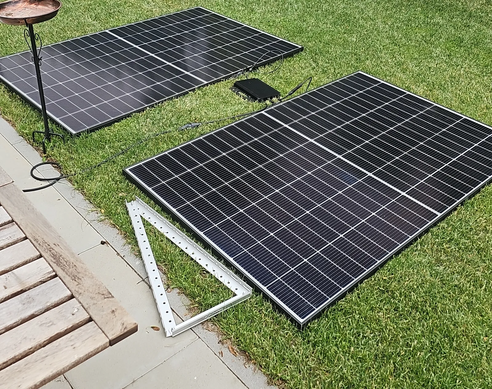 Zwei Solarmodule liegen nebeneinander auf einem Rasen, dazwischen ein kleiner schwarzer Wechselrichter mit Kabeln, davor ein zerlegtes Aluminiumgestell.