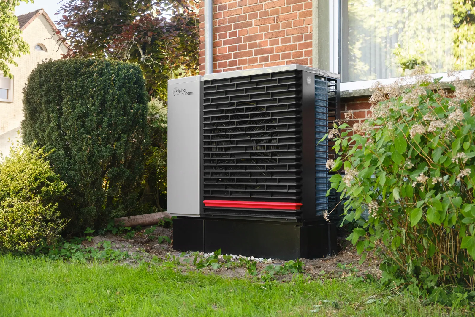 Das Außengerät einer Wärmepumpe steht im Garten vor einem Backsteinhaus, zwischen Büschen.